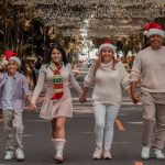 A joyful family walks down a decorated street wearing Christmas hats, symbolizing holiday spirit.
